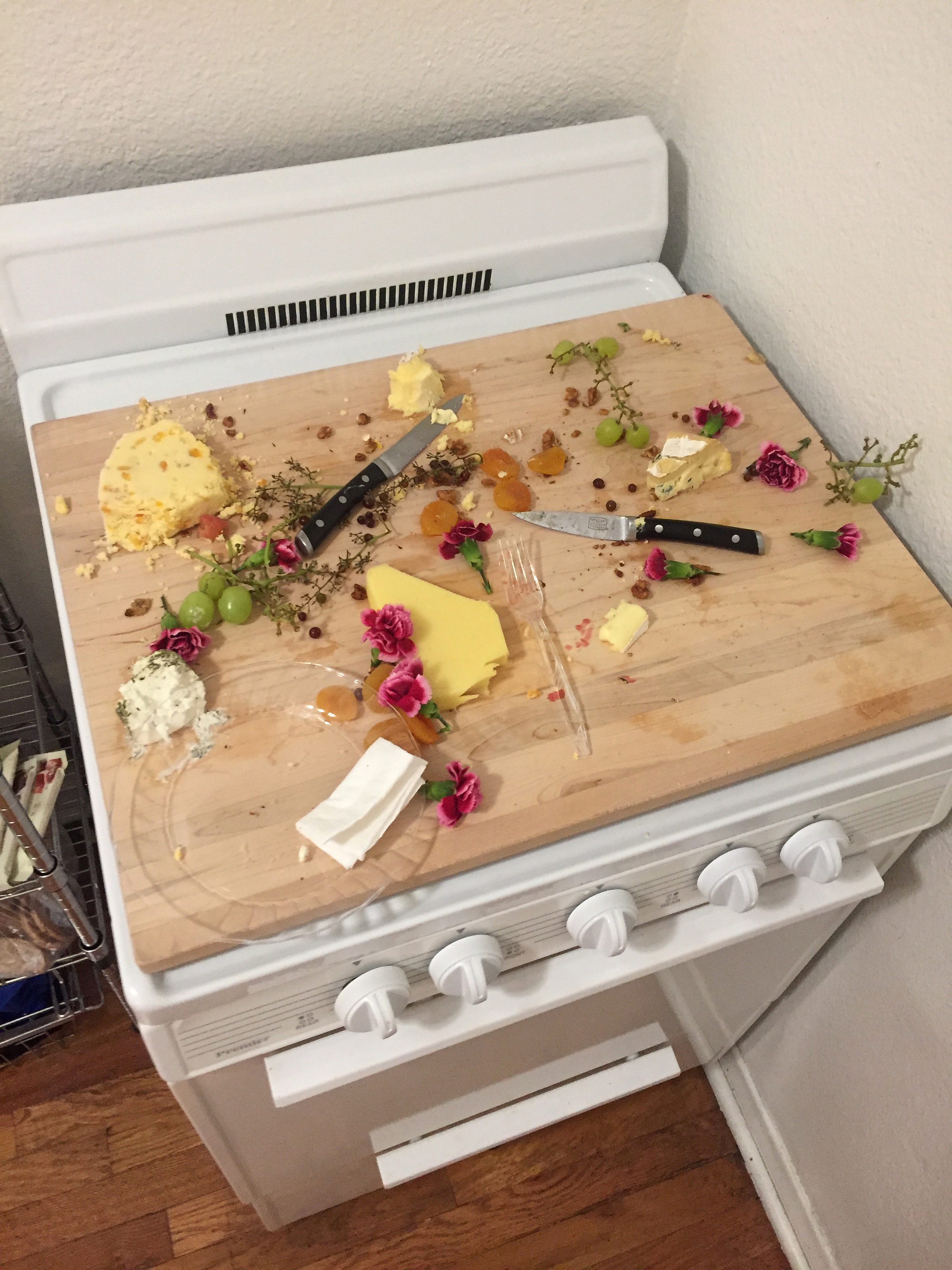 A wooden cheese board littered with food scraps, plates and flowers in a kitchen